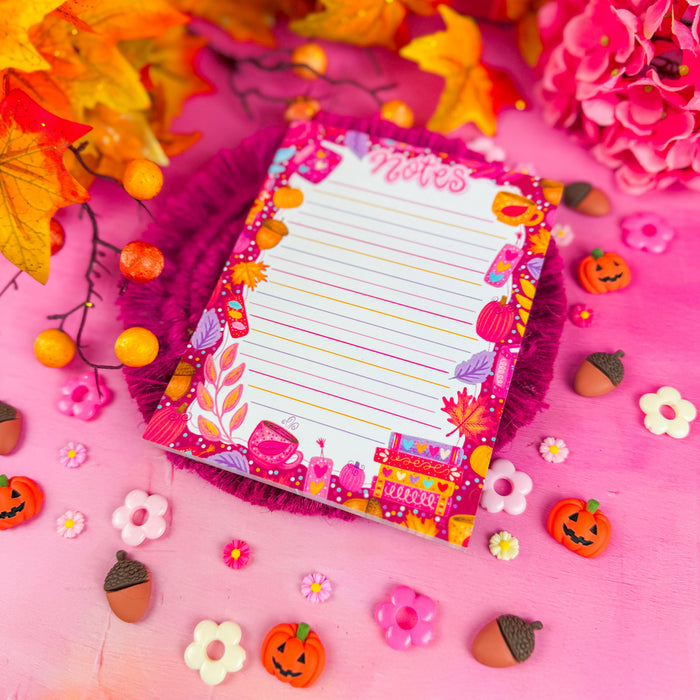 Detail shot of a colorful lined notepad with the word "notes" in pink lettering, and featuring pink and purple leaves, books, and pumpkins around the edges. Against a pink background with pumpkin and flower shapes.