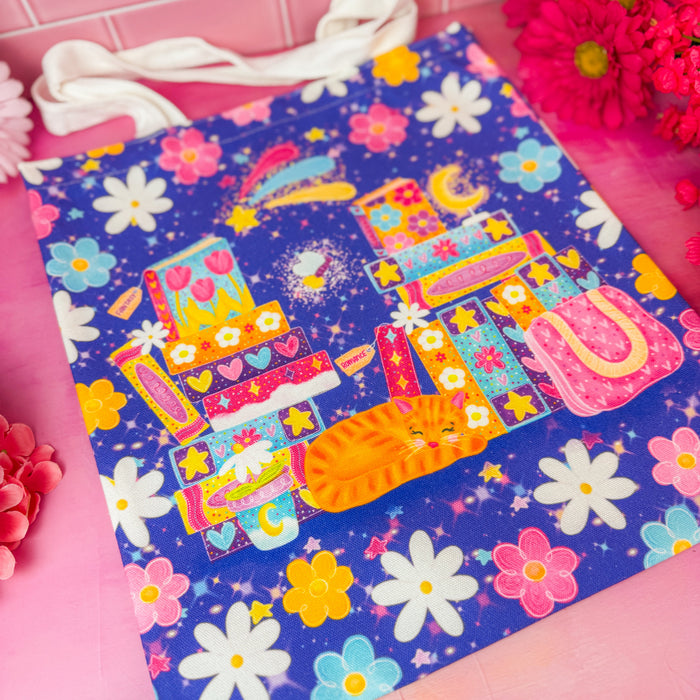 A colorful fabric tote bag with a whimsical illustration of a cat, flowers, and stars on a dark purple background, styled on a pink surface with decorative flowers.