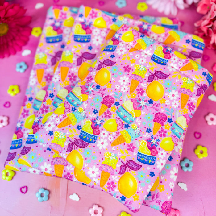 A stack of colorful patterned fabric book sleeves with a playful ice cream and lemon print, arranged on a bright pink surface with decorative flowers and small flower-shaped confetti around them.