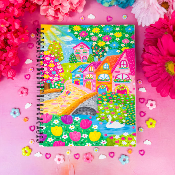A spiral-bound notebook featuring a colorful, whimsical hand-drawn landscape with houses, a bridge, trees, and a swan, styled on a pink background with flowers and small decorative hearts.