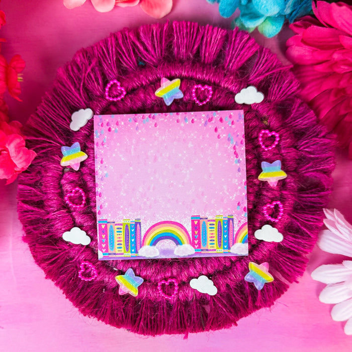 A square pink sticky notepad with a colorful rainbow and book illustration along the bottom edge, placed on a round magenta faux-fur doily decorated with small white clouds, pink hearts, and pastel rainbow star embellishments. Bright pink and blue flowers are blurred in the background.