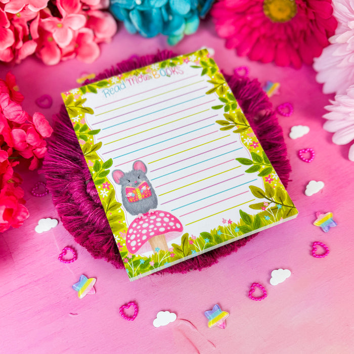 A small lined notepad with a green leaf border, a gray mouse illustration, and a pink mushroom graphic, placed on a pink fringed coaster over a pink surface surrounded by colorful flowers.