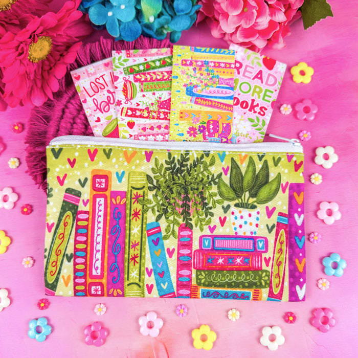 A colorful zippered pouch decorated with a whimsical book-and-plant illustration, shown on a bright pink background with flowers and small flower embellishments. Several matching book-themed cards or envelopes are fanned out from the top.