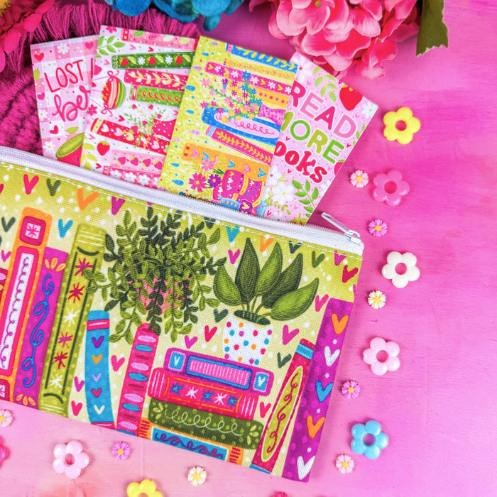 A colorful zippered pouch with a book-and-plant illustration pattern, shown on a bright pink background with flowers and small decorative flowers around it. Several matching patterned paper items are partially visible inside the pouch.