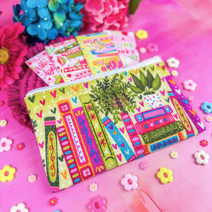 A colorful zippered pouch with a playful book illustration pattern, displayed on a pink wooden surface with decorative flowers and small flower-shaped embellishments.
