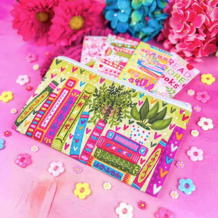 A colorful zippered pouch with a whimsical book-and-plant print, styled on a bright pink surface with flowers and small flower-shaped accessories. A book-themed card is partially tucked into the pouch.