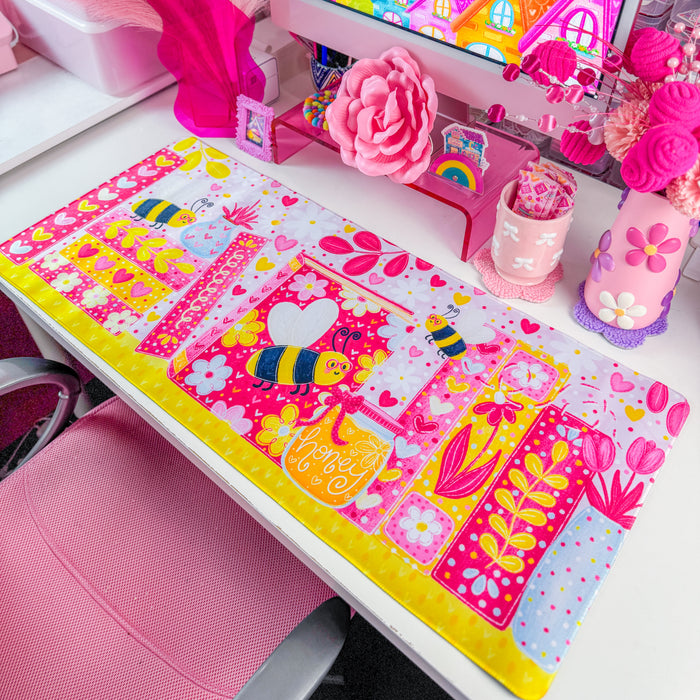 A vibrant pink desk mat with a colorful cartoon bee and floral bookshelf design, styled on a white desk next to a computer monitor in a bright pink room.