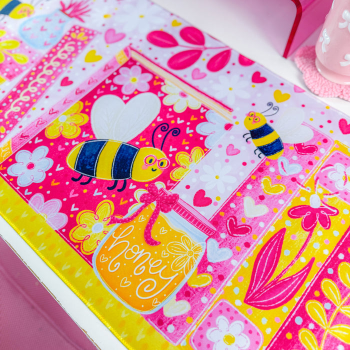 A vibrant pink desk mat with a colorful cartoon bee and floral bookshelf design, styled on a white desk next to a computer monitor in a bright pink room.