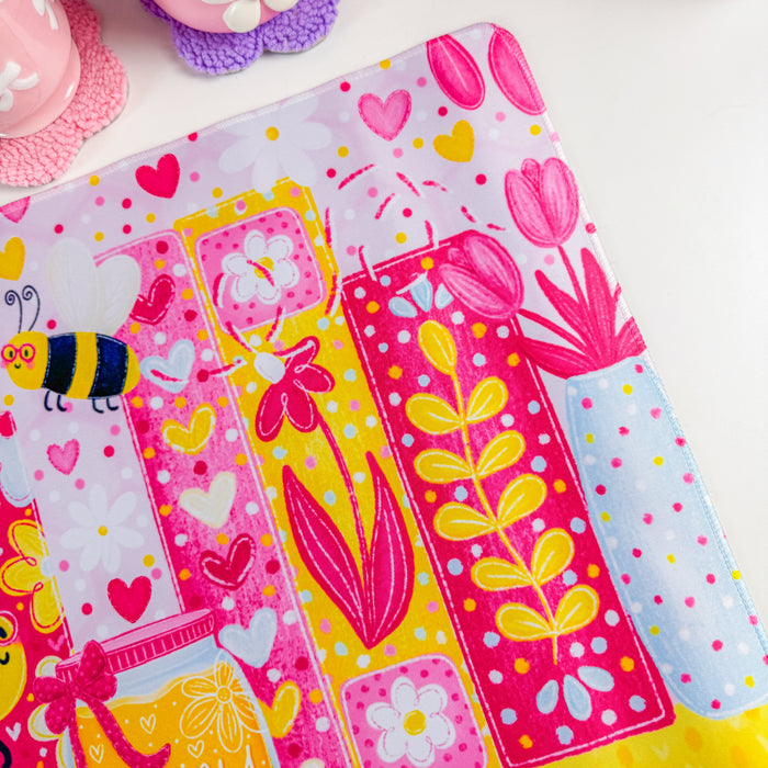 A vibrant pink desk mat with a colorful cartoon bee and floral bookshelf design, styled on a white desk next to a computer monitor in a bright pink room.