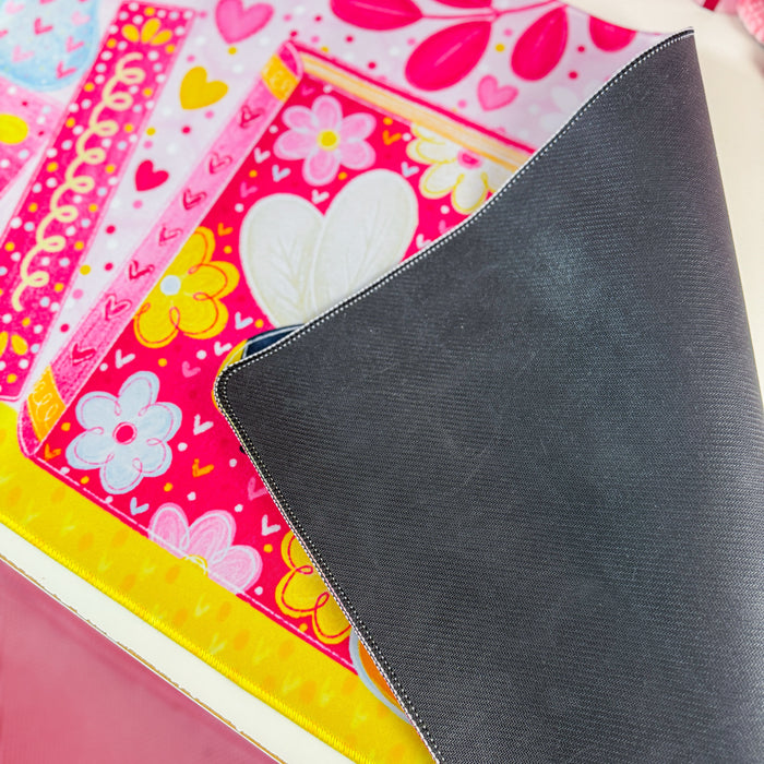 A close-up of a dark gray textured mousepad or desk mat partially covering a colorful patterned desk mat with pink, yellow, and white floral and heart designs.