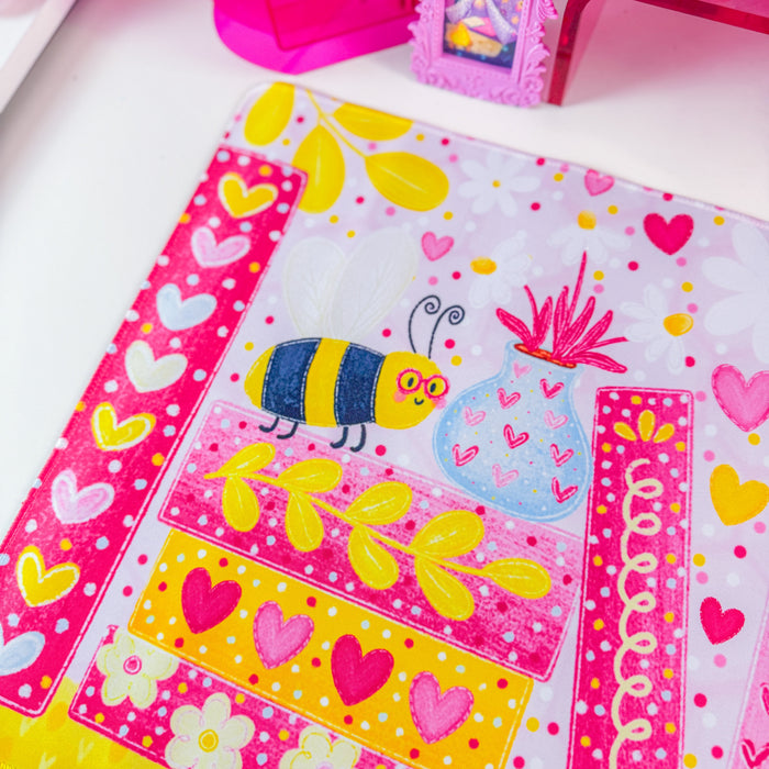 A vibrant pink desk mat with a colorful cartoon bee and floral bookshelf design, styled on a white desk next to a computer monitor in a bright pink room.