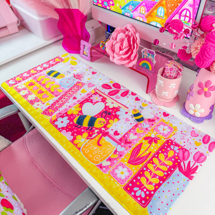 A vibrant pink desk mat with a colorful cartoon bee and floral bookshelf design, styled on a white desk next to a computer monitor in a bright pink room.
