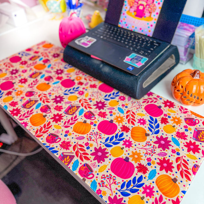 A colorful desk mat with a vibrant autumn pattern of pumpkins, flowers, and leaves, styled on a desk with a laptop and small decorative pumpkins.