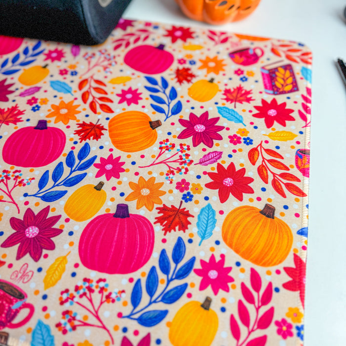A close-up of a fabric or paper product printed with a colorful autumn pattern featuring pumpkins, flowers, and leaves on a light background. The product is photographed on a white surface with partial props visible, including a black device and an orange decorative pumpkin.
