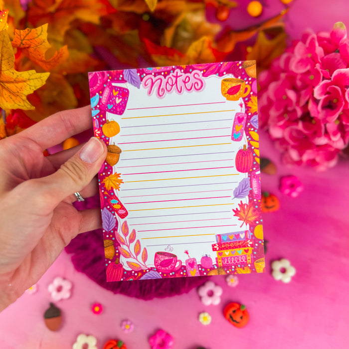 A colorful lined notepad with the word "notes" in pink lettering, and featuring pink and purple leaves, books, and pumpkins around the edges. Against a pink background with pumpkin and flower shapes.