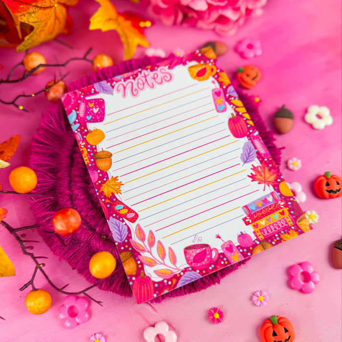 A colorful lined notepad with the word "notes" in pink lettering, and featuring pink and purple leaves, books, and pumpkins around the edges. Against a pink background with pumpkin and flower shapes.