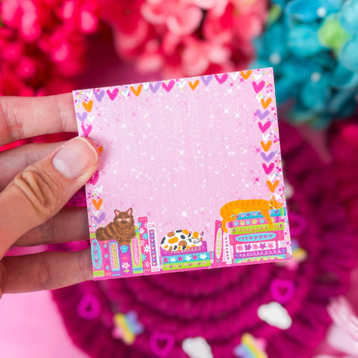 A hand holds a small square pink notepad with a colorful bookshelf illustration and heart border, set against a bright pink floral background.
