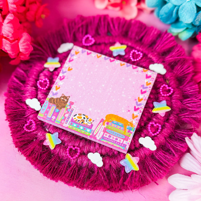 A square pink sticky notepad with a colorful cartoon illustration and heart border, placed on a round magenta fringed coaster surrounded by decorative flowers and small stickers.