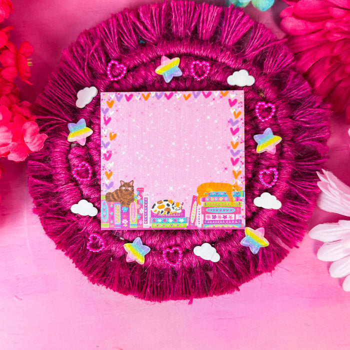 A square pink sticky notepad with a colorful cartoon illustration and heart border, placed on a round magenta fringed coaster surrounded by decorative flowers and small stickers.