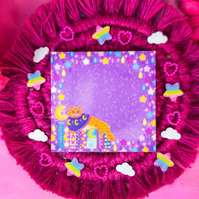 A square sticky notepad featuring a whimsical purple background with a smiling cat under a crescent moon sitting on a book stack, surrounded by a starry border. The notepad is displayed on a round, fringed magenta fabric cushion decorated with small pastel stickers, set against a bright pink background with colorful flowers.