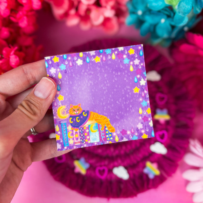 A hand holds a small square sticky notepad featuring a colorful illustration of a cat in a sweater and stars, set against a bright pink background with decorative flowers.