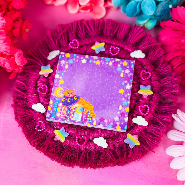 A square sticky notepad featuring a whimsical purple background with a smiling cat under a crescent moon sitting on a book stack, surrounded by a starry border. The notepad is displayed on a round, fringed magenta fabric cushion decorated with small pastel stickers, set against a bright pink background with colorful flowers.