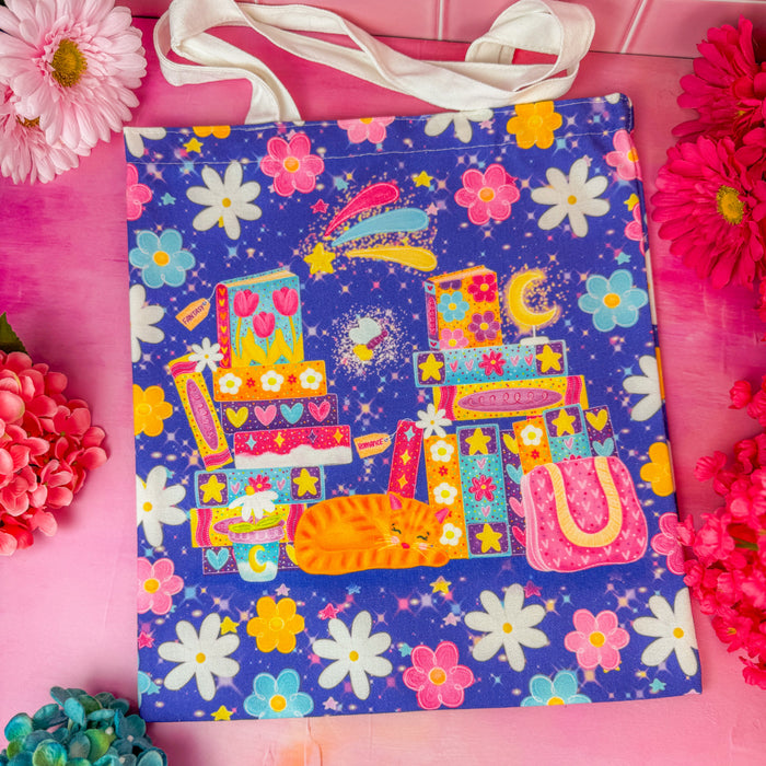 A colorful tote bag with white canvas handles, featuring a vibrant pink, yellow, and blue graphic of stacked books, a crescent moon, and a cat, all over a dark purple floral background. The bag is styled on a pink surface with decorative flowers.