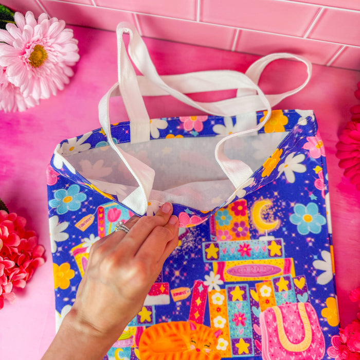 A hand holding open a bright purple tote bag with a colorful print of flowers, books, and small illustrations, set against a pink tiled background with decorative flowers.