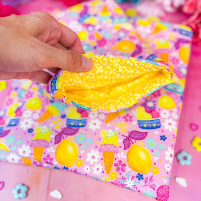 A hand holds open a small book sleeve with a bright yellow interior, placed over a pink patterned fabric book sleeve decorated with colorful ice cream and lemon illustrations.