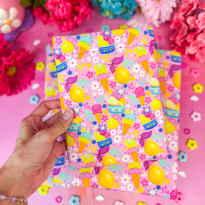 A hand holds a pink book sleeve with a colorful ice cream and candy pattern, surrounded by pink flowers and small flower stickers on a pink background.