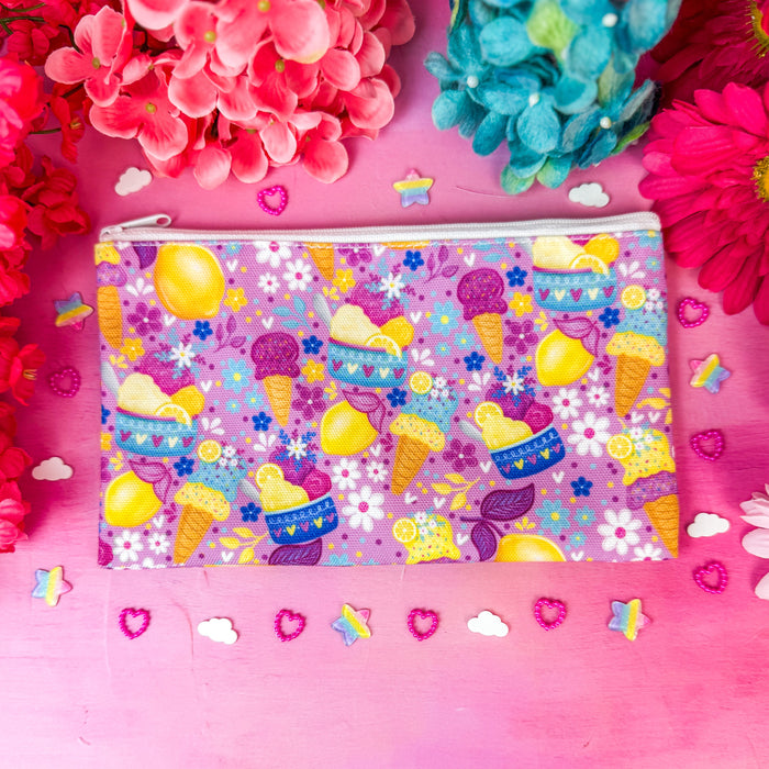 A zippered cosmetic pouch with a colorful pastel pattern of ice cream cones, lemons, and flowers, styled on a pink background with decorative flowers and small confetti.
