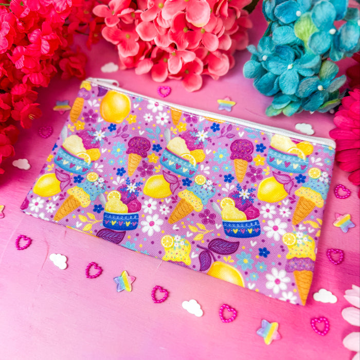 A zippered cosmetic pouch with a colorful pastel pattern of ice cream cones, lemons, and flowers, styled on a pink background with decorative flowers and small confetti.