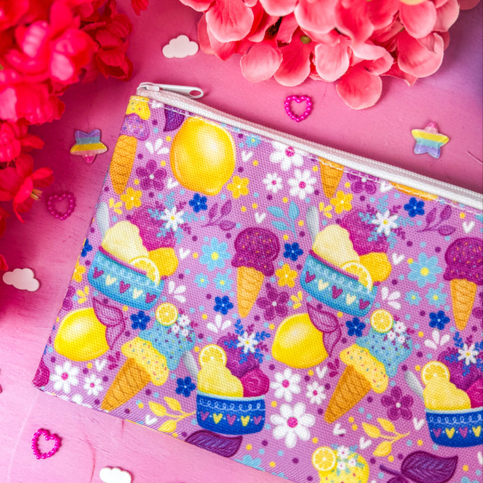 A zippered cosmetic pouch with a colorful pastel pattern of ice cream cones, lemons, and flowers, styled on a pink background with decorative flowers and small confetti.
