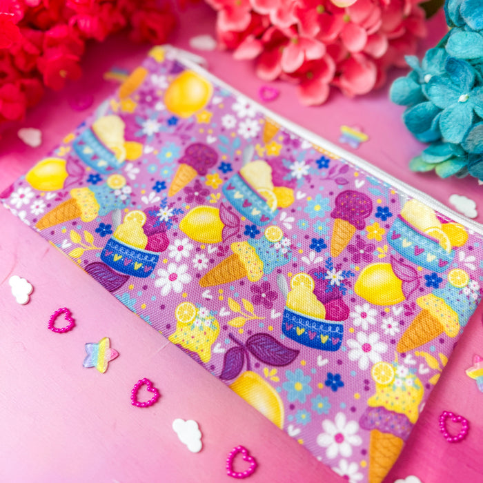 A zippered cosmetic pouch with a colorful pastel pattern of ice cream cones, lemons, and flowers, styled on a pink background with decorative flowers and small confetti.
