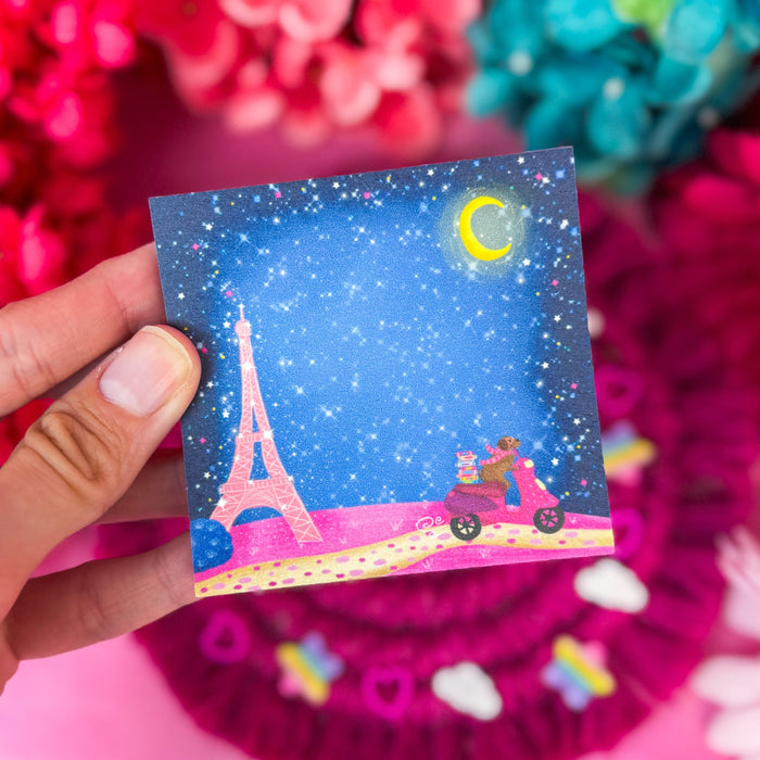 A hand holds a small illustrated sticky notepad featuring a starry blue night sky, Paris, and a pink scooter, with a bright pink floral background.