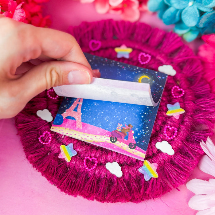 A hand peels back a sheet from a small sticky notepad, placed on a bright pink textured surface with colorful flowers around the edges.