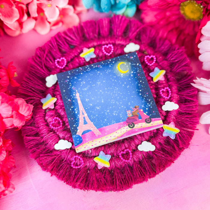 A square sticky notepad featuring a Paris night scene with stars, a moon, and a pink scooter, displayed on a bright magenta fringed fabric pad surrounded by pink and blue flowers.