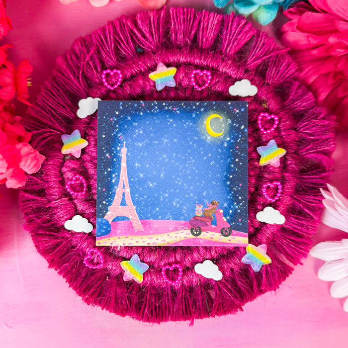A square sticky notepad featuring a Paris night scene with stars, a moon, and a pink scooter, displayed on a bright magenta fringed fabric pad surrounded by pink and blue flowers.