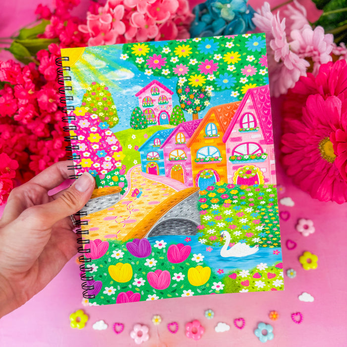 Hand holding a journal featuring a design of a bridge leading through a village up to a bookshop, against a pink backdrop