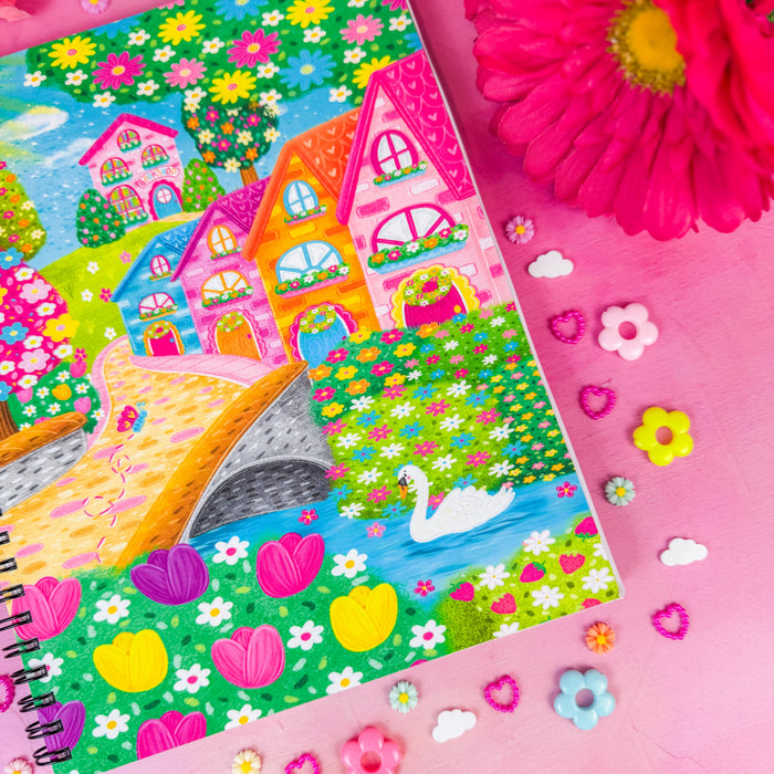 A colorful spiral-bound notebook featuring a whimsical illustration of pastel houses, flowers, a bridge, and a swan, styled on a pink background with decorative flower and heart-shaped accessories.