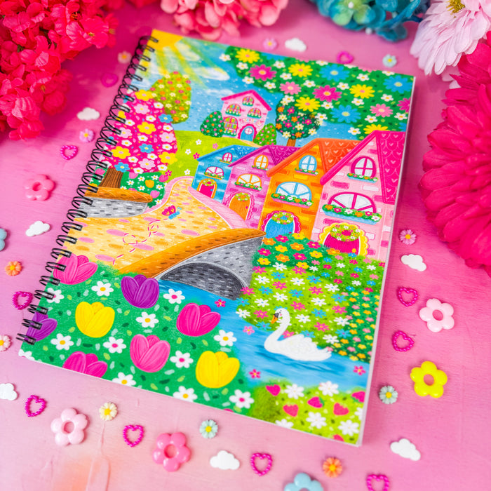 A spiral-bound notebook featuring a colorful, whimsical illustration of houses, a bridge, flowers, and a swan, styled on a pink surface with decorative flower petals and artificial flowers.