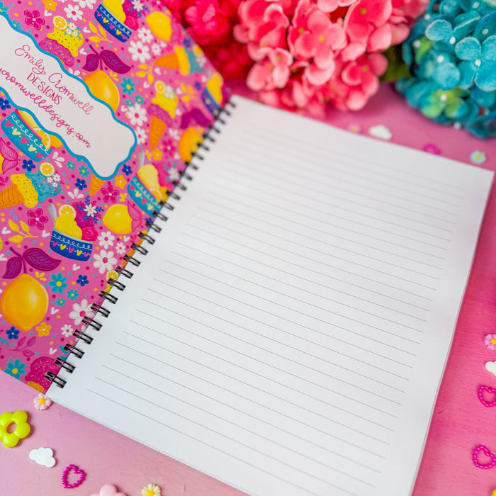 A spiral-bound lined notebook with a colorful floral and lemon-themed interior cover lies open on a pink surface decorated with small flower and heart embellishments. Pink and blue flowers are visible in the background.