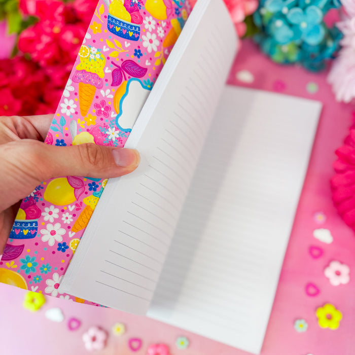 A hand holds an open lined notebook with a bright pink floral-patterned interior cover on a pink surface decorated with small flower and heart embellishments.