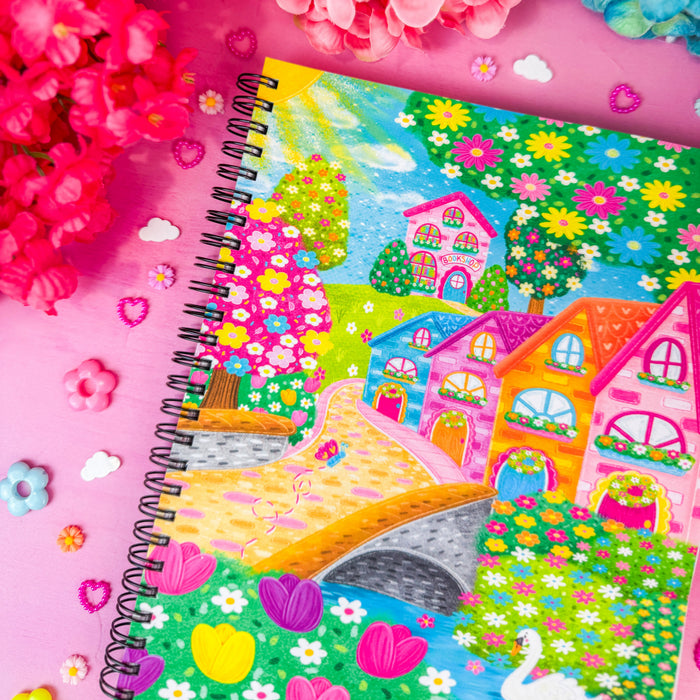 A colorful spiral-bound notebook featuring a whimsical village illustration, styled with bright pink and blue flowers on a pink background.