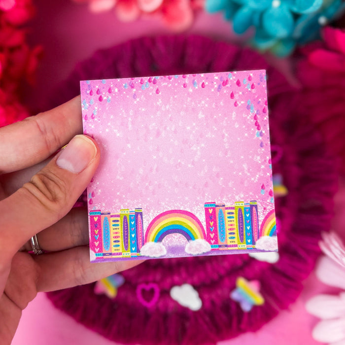 A hand holds a small square pink sticky notepad a colorful rainbow and bookshelf illustration at the bottom, surrounded by bright pink and blue flowers on a pink background.