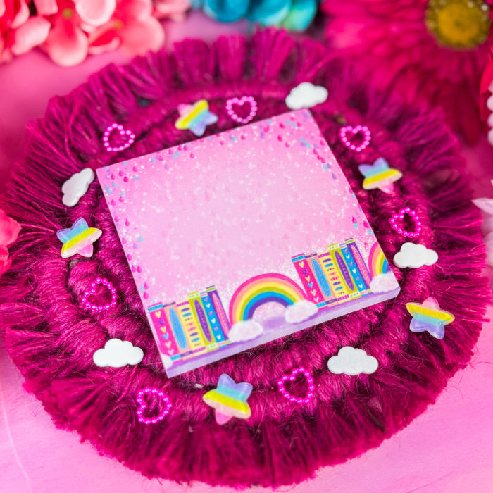 A square pink sticky notepad with a colorful rainbow and book illustration along the bottom edge, placed on a round magenta faux-fur doily decorated with small white clouds, pink hearts, and pastel rainbow star embellishments. Bright pink and blue flowers are blurred in the background.