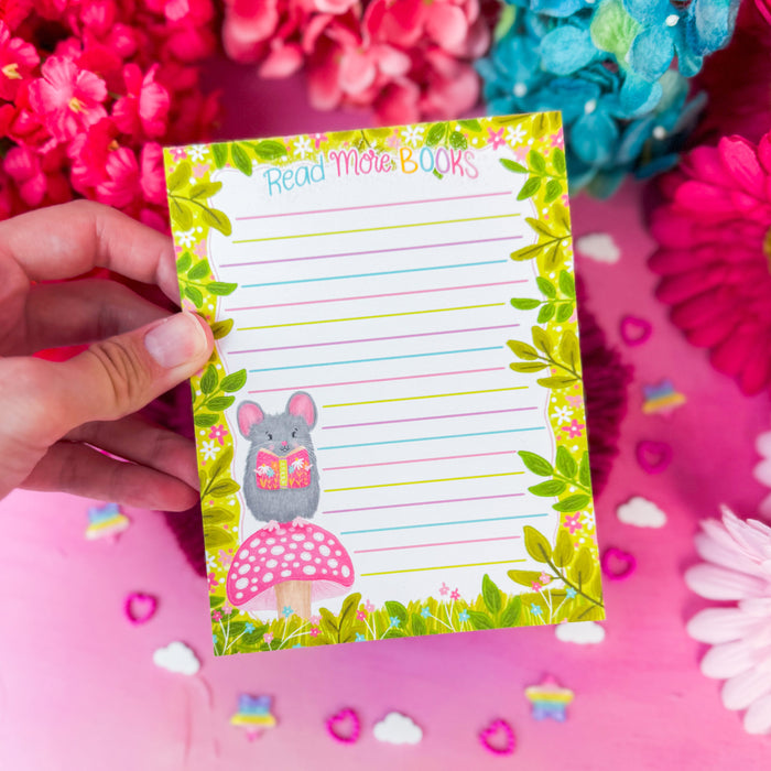 A hand holds a lined notepad featuring a cute mouse reading a book on a mushroom, surrounded by a green leaf border. The notepad is set against a pink background decorated with colorful artificial flowers.