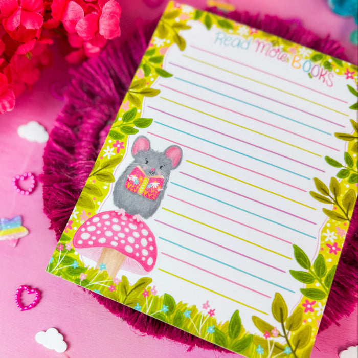 A small lined notepad with a green leaf border, a gray mouse illustration, and a pink mushroom graphic, placed on a pink fringed coaster over a pink surface surrounded by colorful flowers.