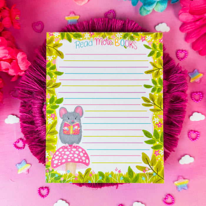 A small lined notepad with a green leaf border, a gray mouse illustration, and a pink mushroom graphic, placed on a pink fringed coaster over a pink surface surrounded by colorful flowers.
