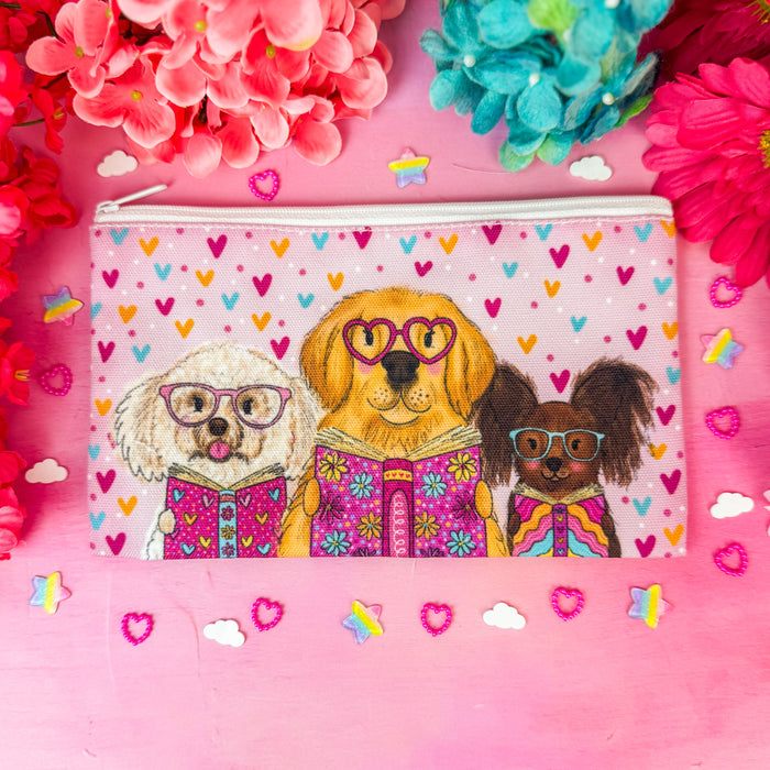 A rectangular zip pouch with a white background and a colorful heart pattern, featuring an illustration of three dogs wearing glasses and colorful patterned sweaters. The pouch is styled in a flat-lay arrangement on a pink surface, surrounded by pink and blue flowers.